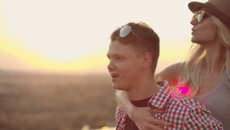 una pareja joven se mueve y gira en un baile al atardecer. una mujer con cabello rubio en un sombrero y gafas está sentada en la espalda de su novio.
