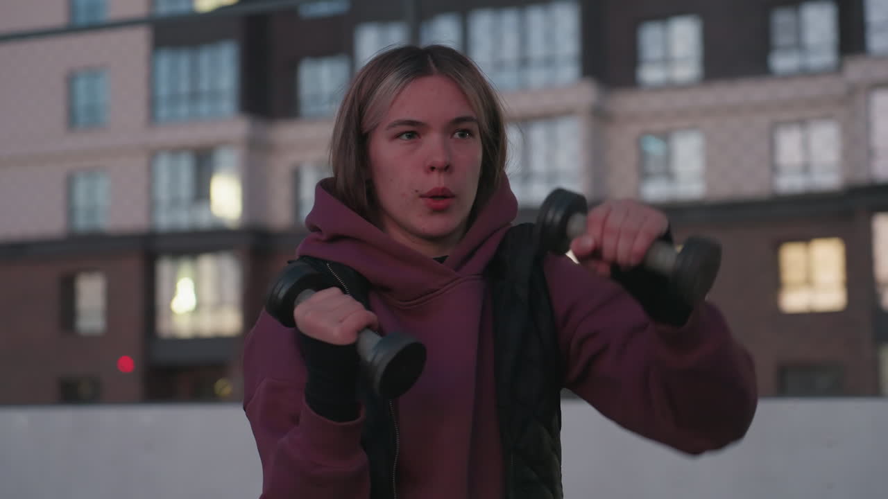 Urban woman boxing dumbbells punching air at dusk on black asphalt sports court next to white barrier topped with chain link fence wearing maroon hoodie and white sneakers with tall building backdrop