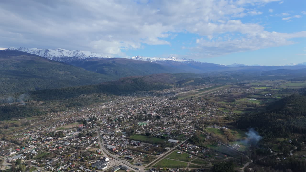 Slow upward tilting wide angle drone shot over El Bolsón town in Argentinean South with houses and buildings at surface and stunning Andean mountain range in background. Shot on DJI Air 3 at 4K-60fps.