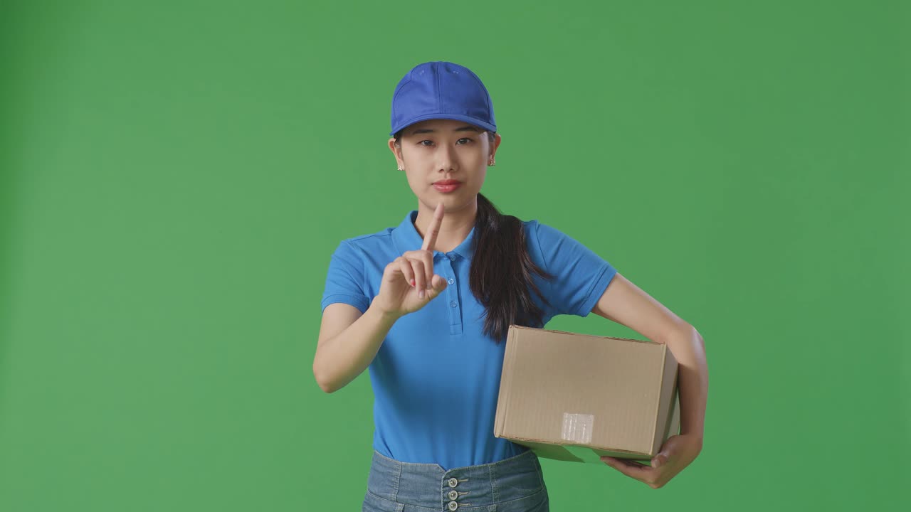 푸른 유니폼을 입은 아시아 여성 택배기사는 스튜디오에서 녹색 스크린 배경에 상자를 배달하는 동안 제스처를 표시하지 않습니다.