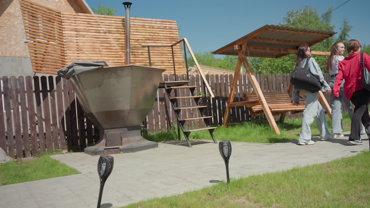 Happy high school students walk out park along paved path, carrying bags and chatting while passing wooden swing and outdoor structure under bright sky in suburban yard with wooden fence