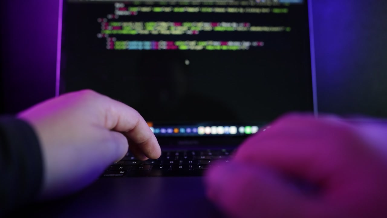 Dark cinematic close-up footage of a person coding on a laptop in a modern neon-lit environment with blue and purple ambient lighting.