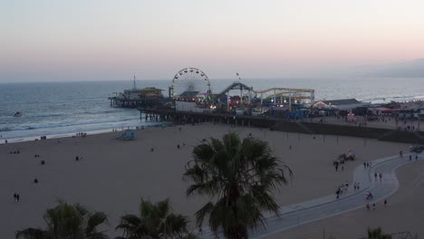 aerial close-up shot flying over palm trees to reveal the santa monica pier on a bustling summer night in los angeles, california at twilight