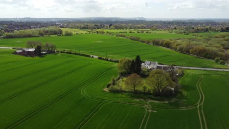 Rural-English-farmhouse-aerial-view-surrounded-by-lush-green-trees-and-agricultural-farmland-countryside-fields