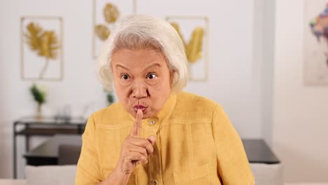 asian grandmother in yellow shirt holds finger to lips, signaling silence with expressive facial gestures in a well-lit, modern home interior. static camera, soft daylight