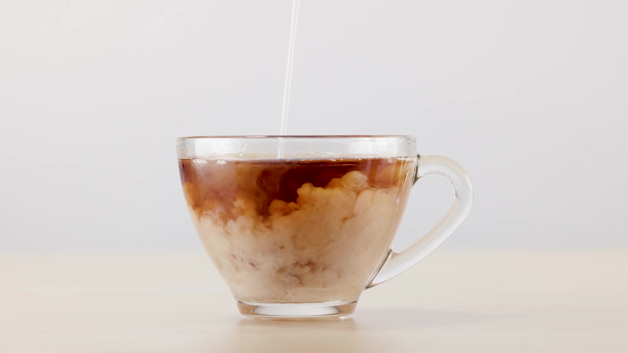 Il latte si versa in una tazza di caffè di vetro trasparente, creando nuvole bianche vorticose contro il marrone.