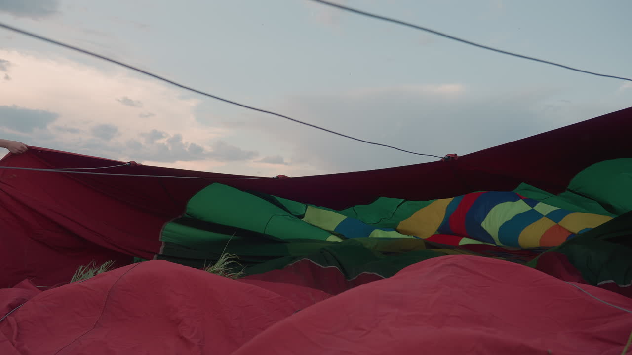 woman and girl holding edges of hot air balloon envelope on grass field under cloudy sunset sky coordinating preflight ground spread of colorful nylon fabric for inflation near trailer