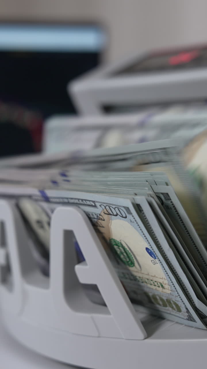 Switching on cash counting machine to count a wad of one hundred dollar bills. Close up. Laptop at backdrop in blur. Vertical video