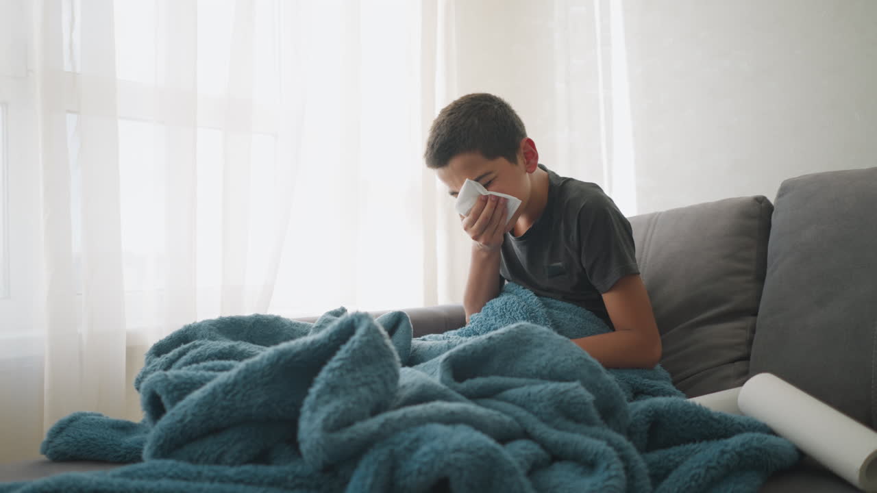 Bambino che starnutisce in un fazzoletto seduto sul divano sotto una coperta blu, con un rotolo di fazzoletti vicino, che mostra segni di malattia, affaticamento e guarigione, che mostra assistenza domiciliare nel soggiorno