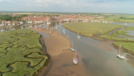 establishing drone shot coming in towards wells-next-the-sea coastal town with salt marsh and creek and sailing boats at low tide in north norfolk uk east coast