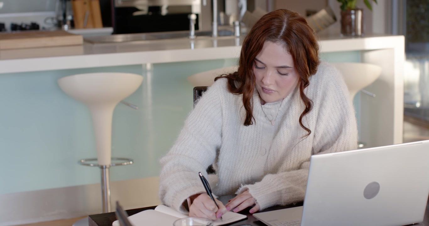 Kvinde i sweater skriver i notesbog ved køkkenbordet, mens hun bruger en bærbar computer til studier.