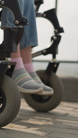 woman pushes medical equipment with schoolgirl after surgery walking in city. legs of little child sitting in wheelchair on walk with mom closeup