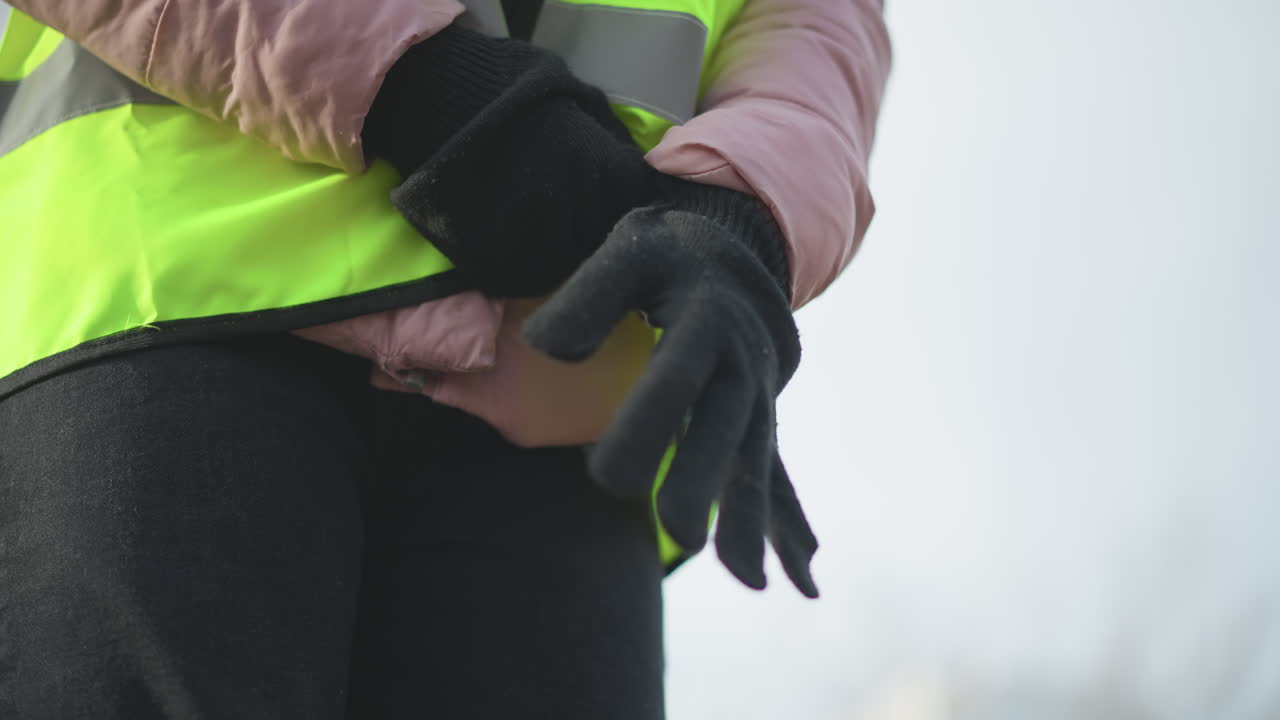 Close-up of person in neon yellow high-visibility vest with reflective stripes and pink padded jacket putting on black knitted gloves outdoors, preparing for safety or work duties in cold winter environment