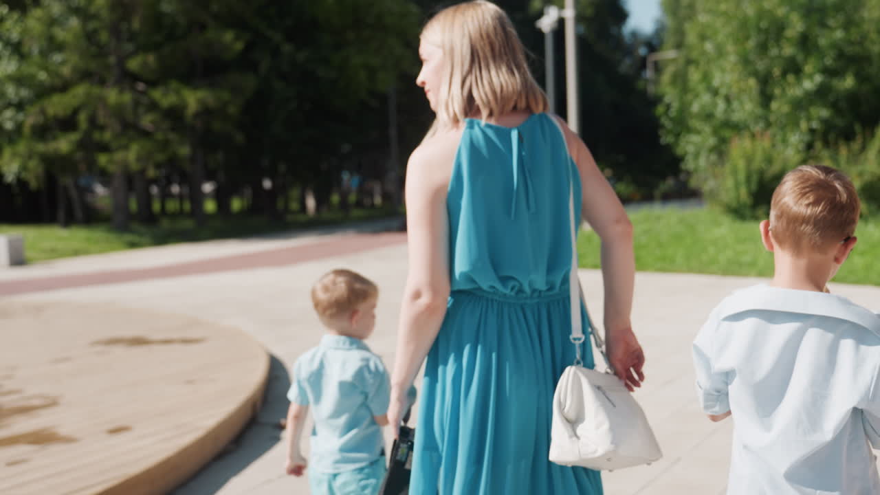 Familie loopt langs een fontein en eet een ijshoorntje, moeder en twee jongens wandelen over het plein bij waterstralen in een blauwe jurk en zomerse outfits, zonlicht en groen, ontspannen samenzijn tijdens een weekenduitje in het park.