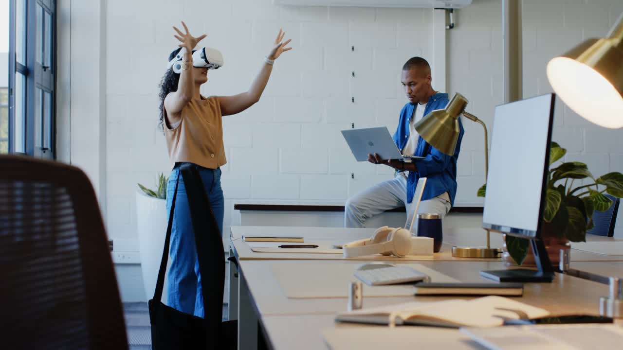 Colleagues at desk, woman initiating VR gestures with headset, guiding man using laptop