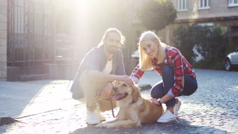 Young-Couple-Crouching-On-The-Street-And-Petting-Their-Corgi-Dog-While-Looking-At-Camera-On-A-Sunny-Day