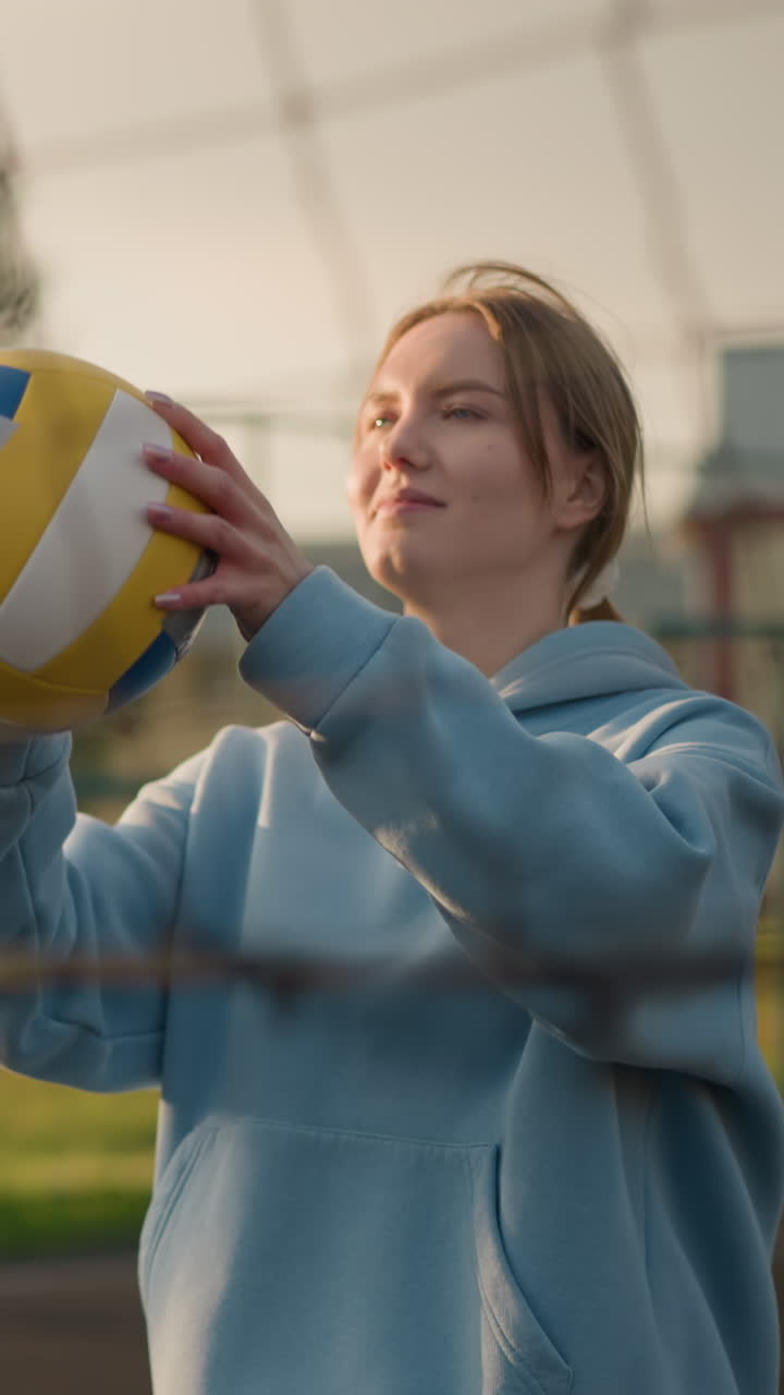 야외 코트에서 볼리볼을 하는 후디를 입은 젊은 여성, 행동에 집중하고, 볼리폴을 들고, 전면에 네트가 보이고, 배경에는 흐릿한 초록색과 건물이 있습니다.