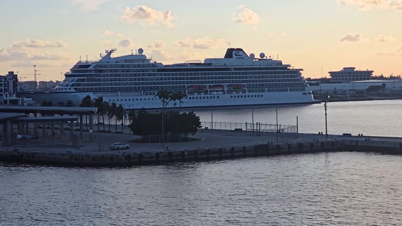 Grande nave da crociera di lusso ormeggiata a un terminal portuale commerciale al tramonto. Acque calme e palme circondano il molo di cemento sotto un cielo luminoso all'ora d'oro.