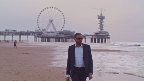 Un-Hombre-Modelo-Latino-Negro-Con-Traje-Camina-Por-La-Playa-Con-Gafas-De-Sol-En-Los-Países-Bajos,-La-Haya