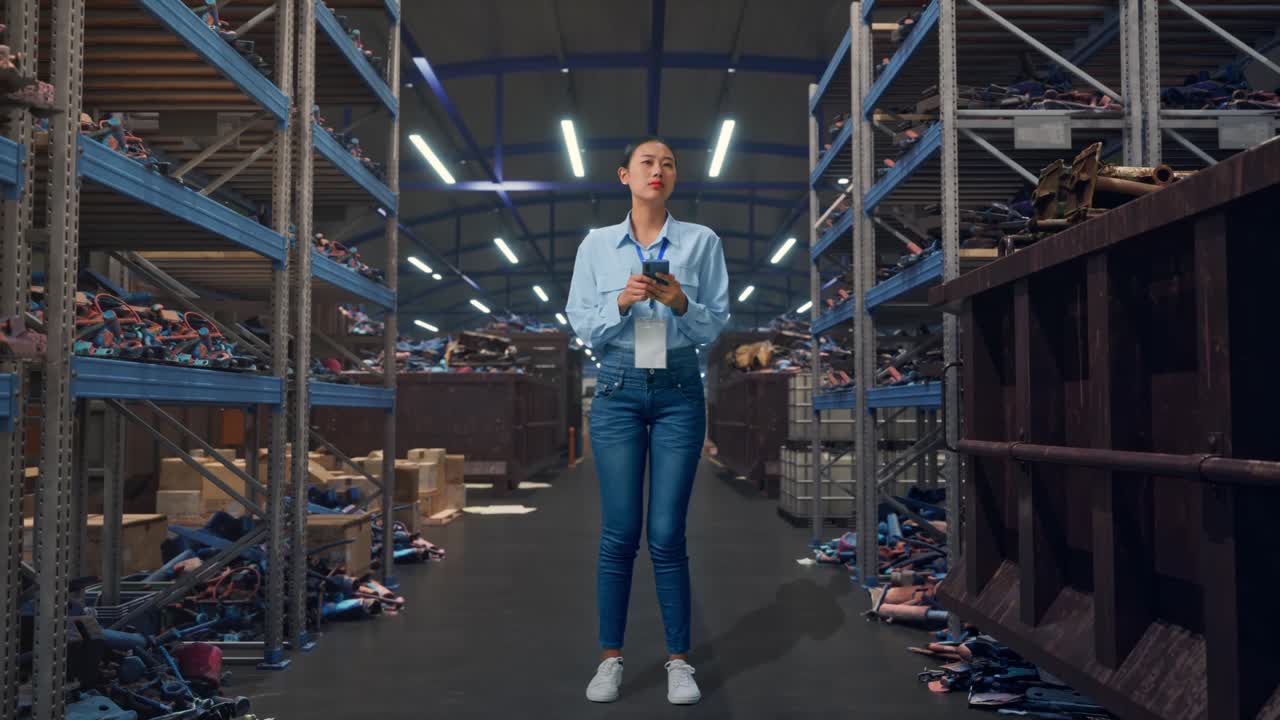 Trabalhadora asiática de corpo inteiro com seu smartphone, em um armazém industrial de sucata e reciclagem. Ela observa ao redor antes de se concentrar em seu smartphone.