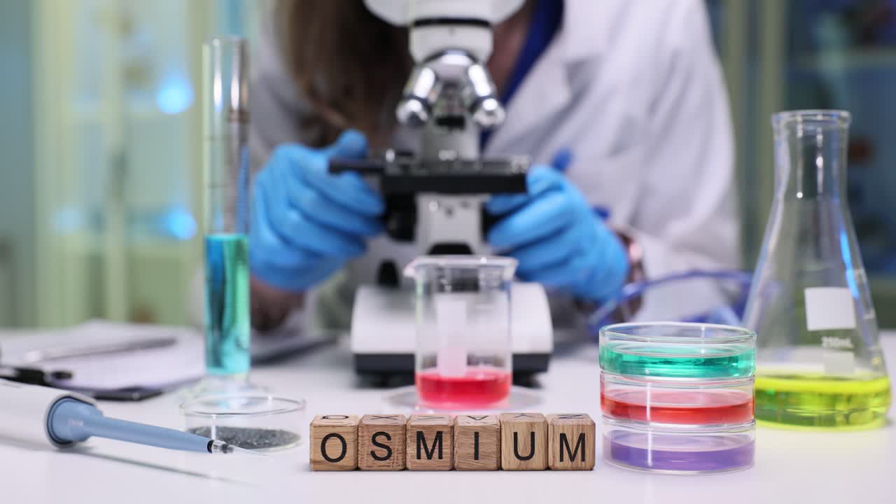 A scientist working with a microscope and various chemicals in a laboratory, with "OSMIUM" spelled out in wooden blocks on the table