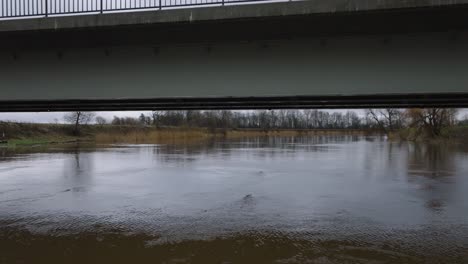 aerial establishing view of high water in springtime, barta river flood, brown and muddy water, overcast day, low drone shot moving forward below the concrete bridge