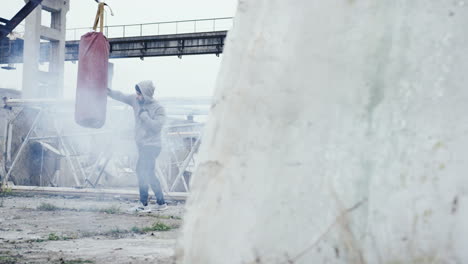 distant view of caucasian man in grey hoodie hitting a punching bag outdoors an abandoned factory on a cloudy morning