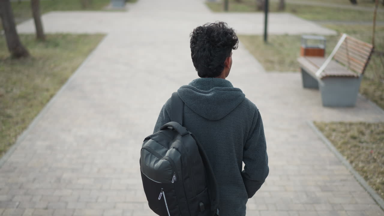 Young man in gray hoodie walking alone down paved park path, seen from behind, carrying black backpack, surrounded by leafless trees and empty benches on overcast day, suggesting solitude or reflection