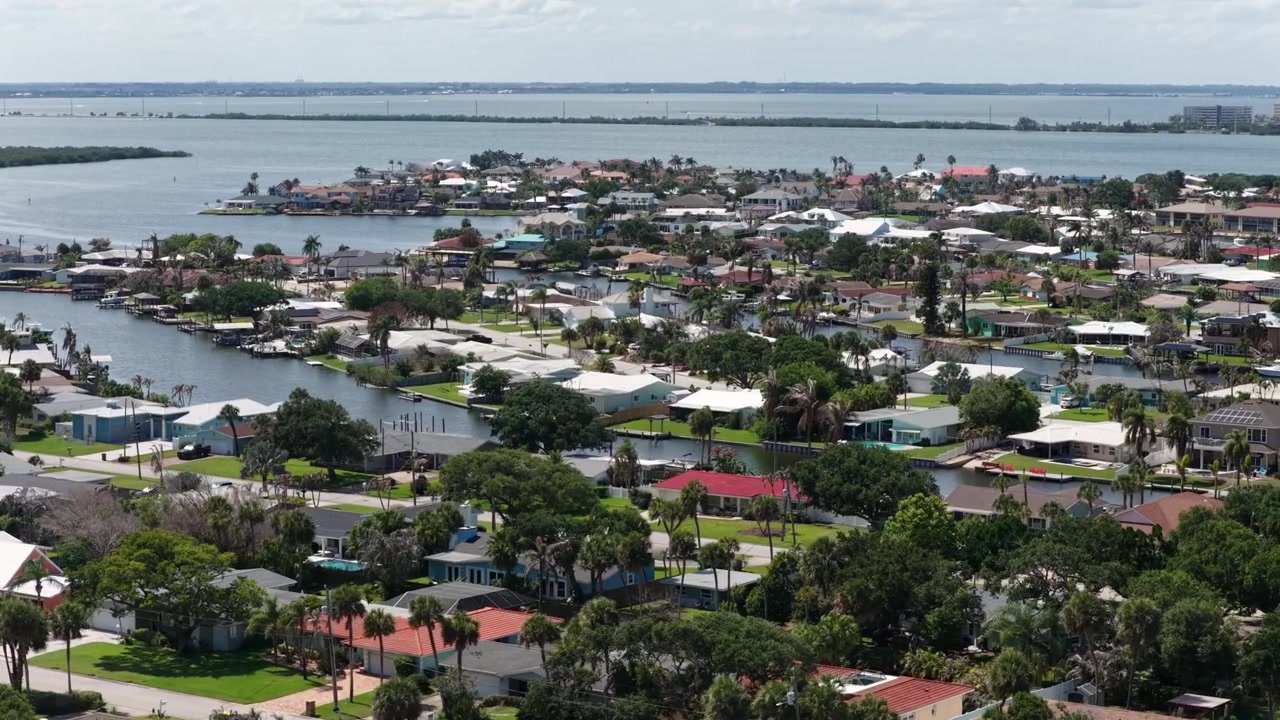 Case sul lungomare con pontili privati costeggiano un quartiere con canali tortuosi a Cocoa Beach, Florida, con palme e tetti in tegole rosse sparsi sulla penisola verso la baia aperta. Ampia ripresa aerea con drone.