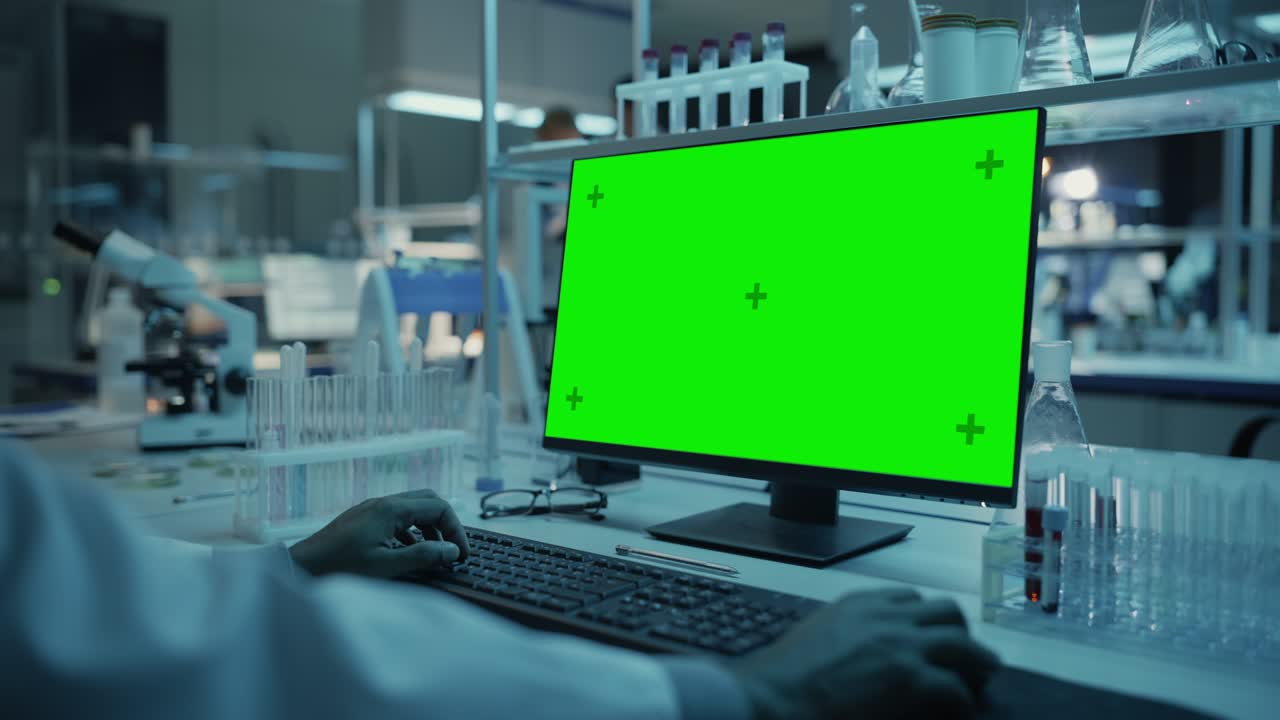 응용 과학 연구 실험실에서 녹색 화면 모형 템플릿을 가진 데스크 컴퓨터에서 일하는 의료 연구 과학자.  코트를 입은 실험실 엔지니어가 배경에서 실험을 수행합니다.