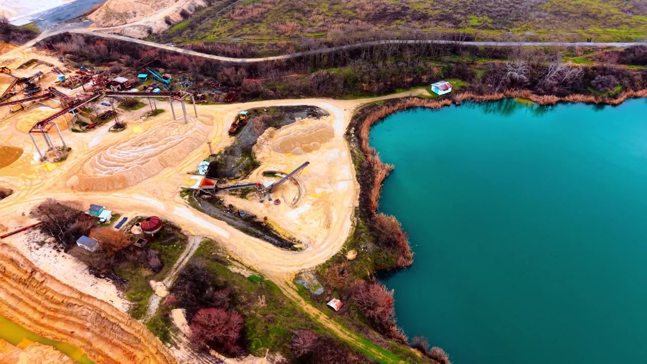 Elevated view of sand processing plant beside turquoise quarry lake. Oblique drone angle showing conveyor towers, sand piles, reed-lined shore and vivid teal water under winter light.