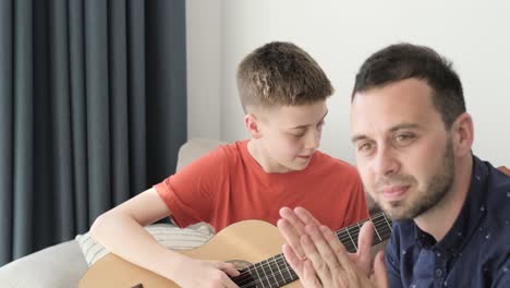a teenage boy plays the guitar in the room with his dad. dad teaches his son to play the guitar. family time together