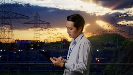 side view of an asian male professional worker standing with his tablet near high voltage tower, industrial facility, checking on his tablet with meditation
