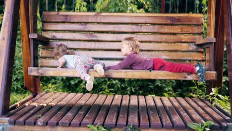 children swing on a large wooden swing near the summer house they play against the background of green trees