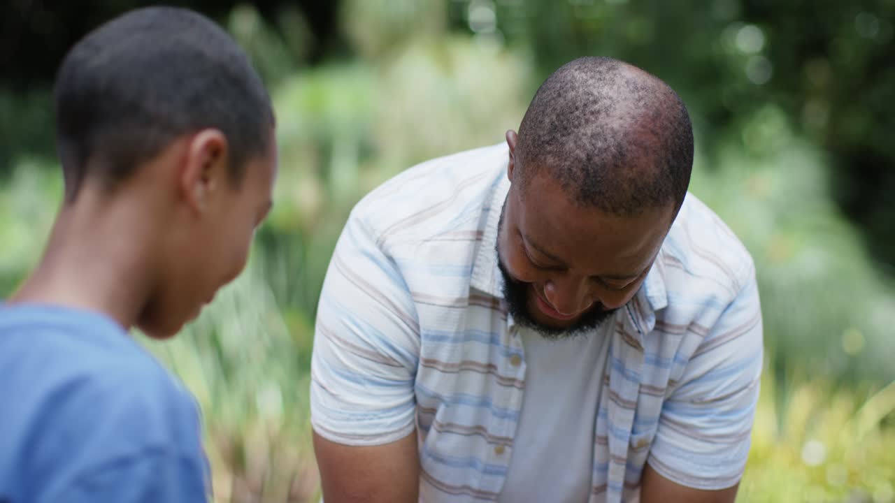 Pai afro-americano com o filho oferecendo um tubérculo roxo; o pai, de camisa listrada, inspecionando no jardim.