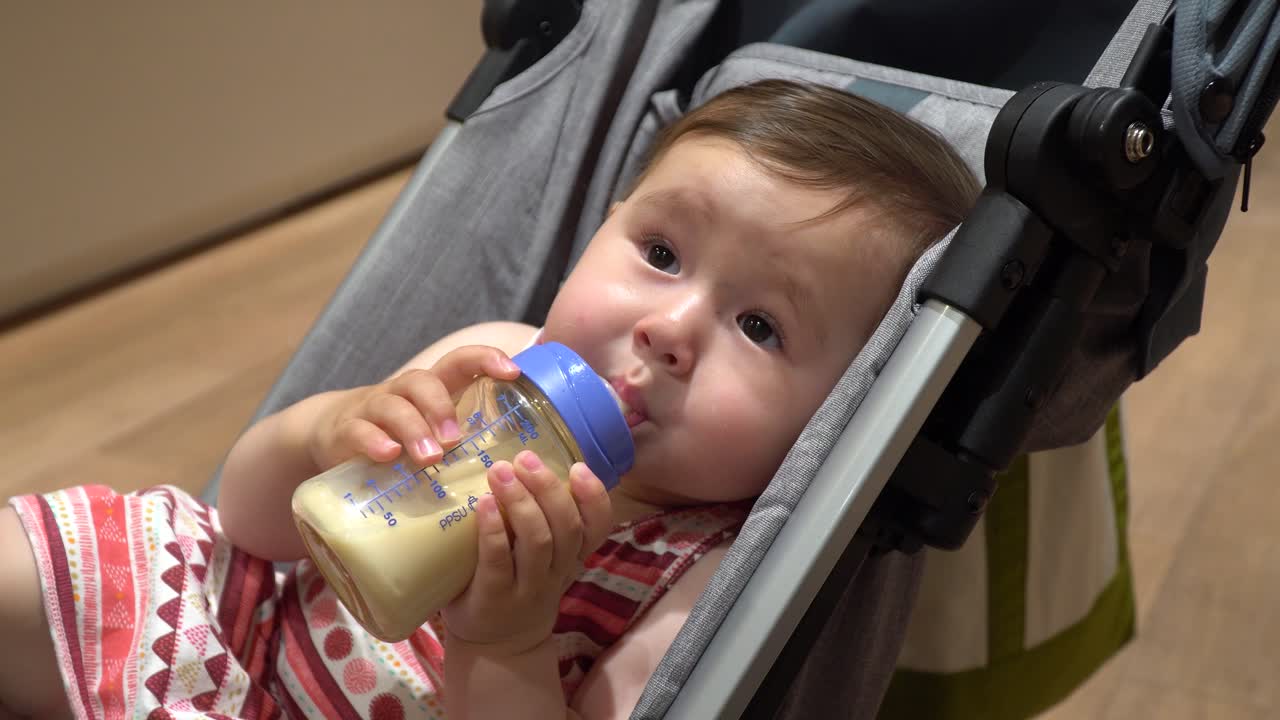 Una tenera bambina di un anno che beve latte dal biberon, tenendolo con entrambe le mani mentre è sdraiata in un passeggino e si guarda intorno - primo piano, inquadratura statica.