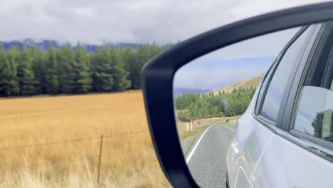 a vehicle travels along a winding rural road, captured through the side mirror with golden fields, green trees, and soft daylight. smooth camera movement enhances the tranquil atmosphere