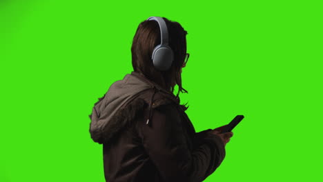 vista trasera de una mujer joven con auriculares inalámbricos transmitiendo música desde un teléfono móvil contra la pantalla verde del estudio