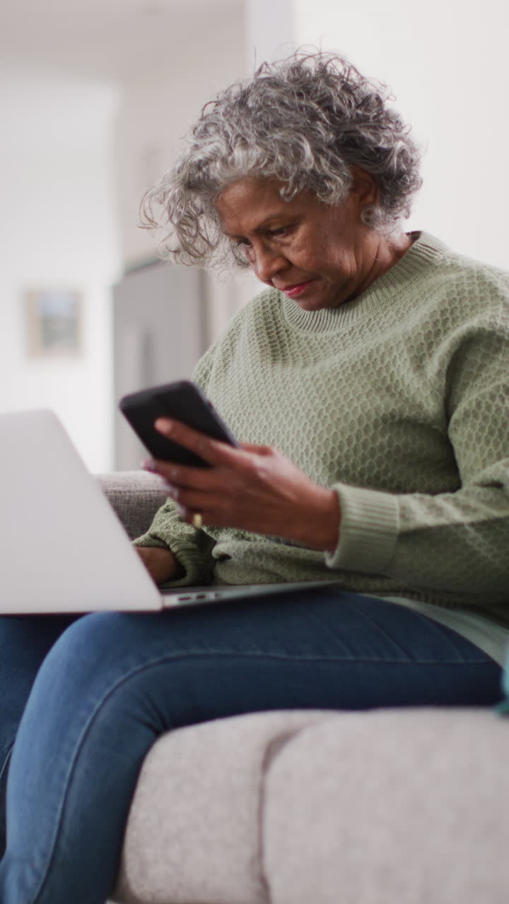 Vertical video of happy senior african american woman sitting on sofa, using laptop and smartphone