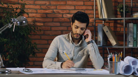 joven arquitecto hablando por teléfono alegremente mientras se sienta en el escritorio dibujando en él