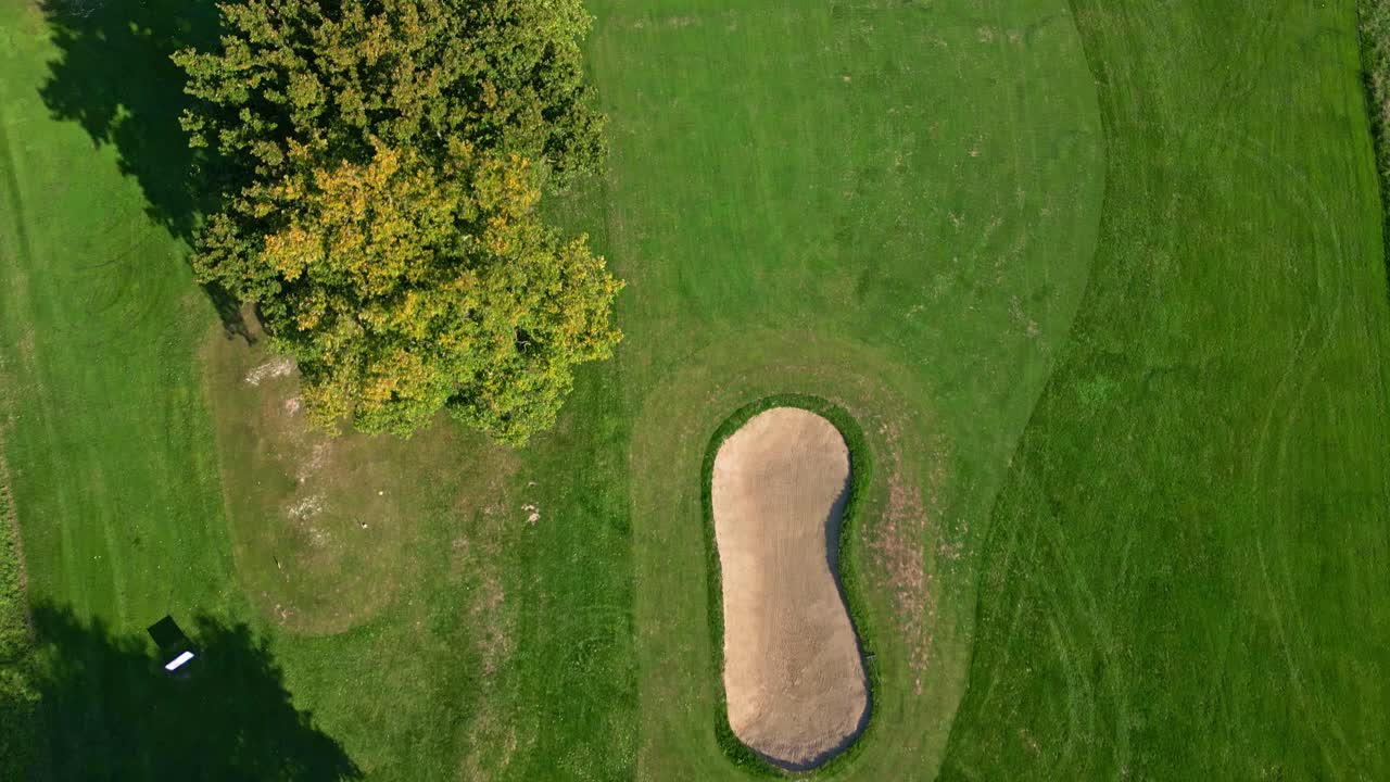 Drone top-down shot moving over a golf course in France, showing sand bunker, green grass, trees, and a winding path under sunlight