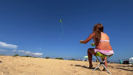 Niña-Pelirroja-Sentada-En-Una-Pequeña-Silla-De-Playa-Disfruta-Volando-Una-Cometa-De-Doble-Hebra-En-Un-Caluroso-Día-De-Verano