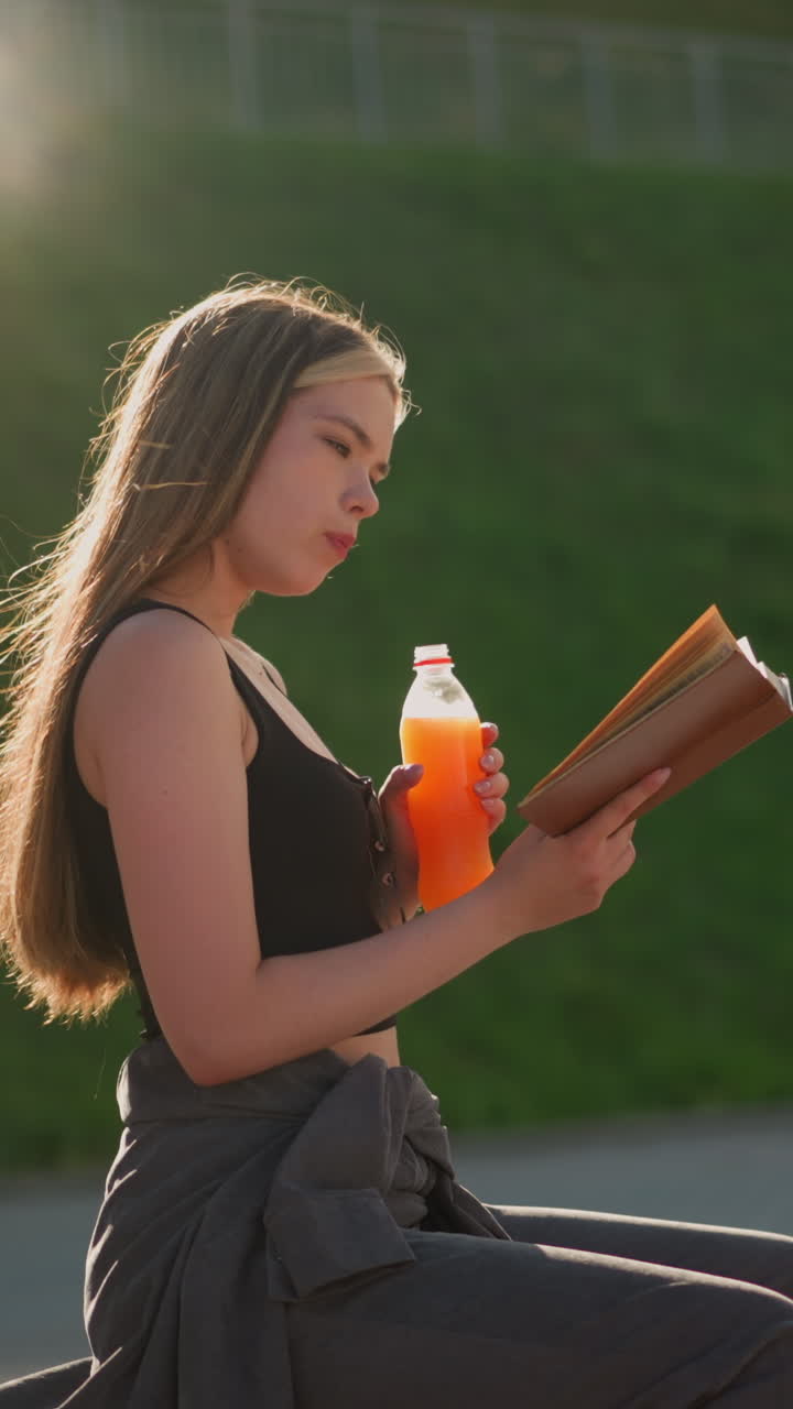 mujer sentada al aire libre, sosteniendo un libro en una mano y bebiendo de una botella de jugo con la otra, continúa leyendo mientras la luz del sol crea un ambiente cálido en el fondo