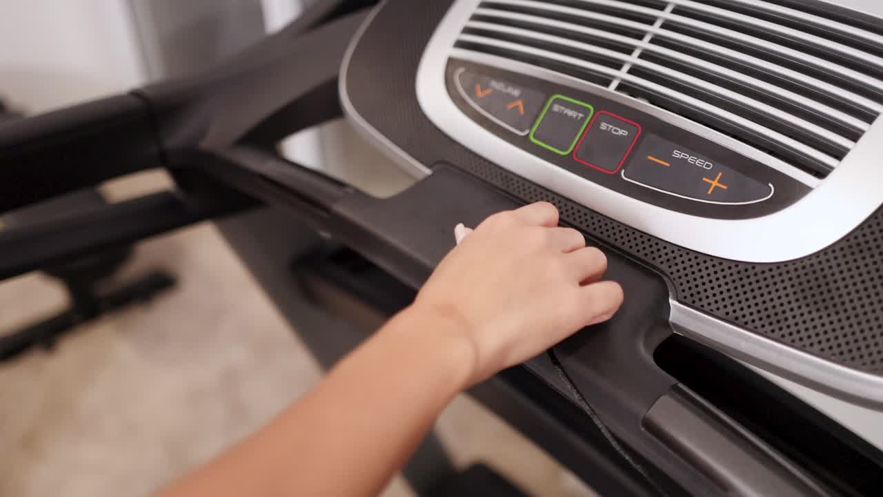 A woman interacts with a treadmill control panel in a home gym, adjusting settings under warm indoor lighting