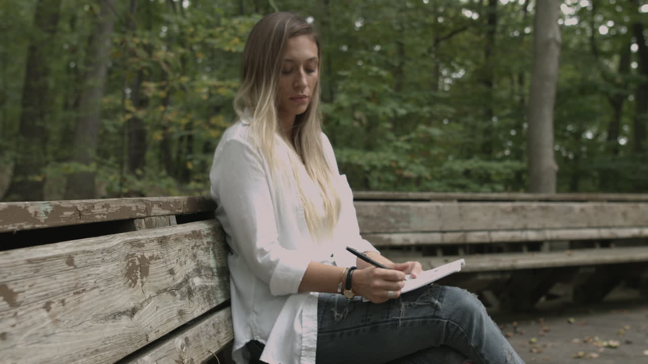 Still shot blonde female girl sits in the woods with a note pad drawing taking notes