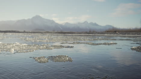 Ice-flows-by-in-an-Alaska-river,-with-huge-snow-capped-peaks-in-the-distance