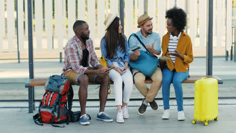 multiethnic group of travellers sitting at bus stop talking while holding tablet and maps