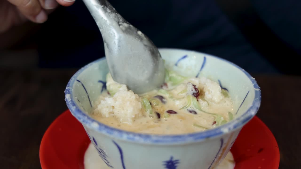 Hand uses a metal spoon to scoop cendol, shaved ice, and red beans from a patterned ceramic bowl under soft indoor lighting, close-up perspective