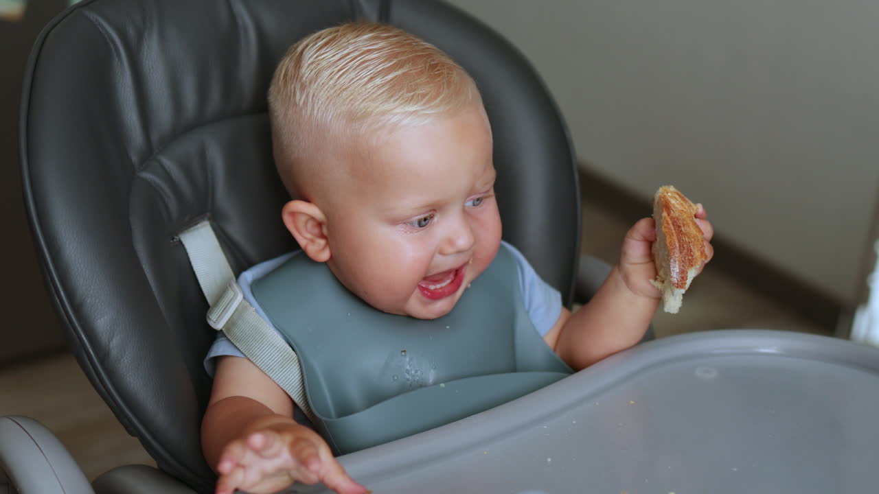 One-year-old boy sits in a high chair and learns to eat by himself. Silicone tableware for children. Boy eats from a plate by himself