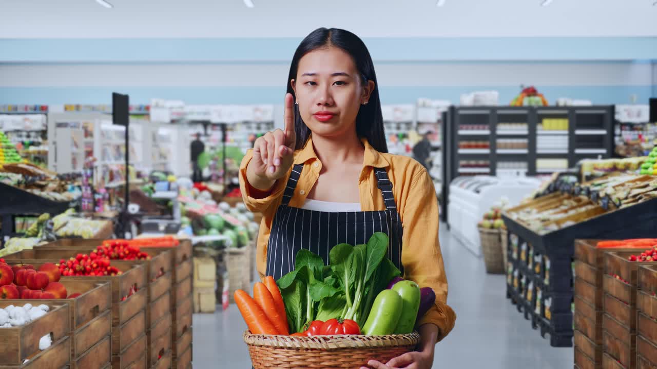 Agricultora asiática con cesta de verduras mostrando el gesto de parar con un dedo, señal de tabú en un supermercado